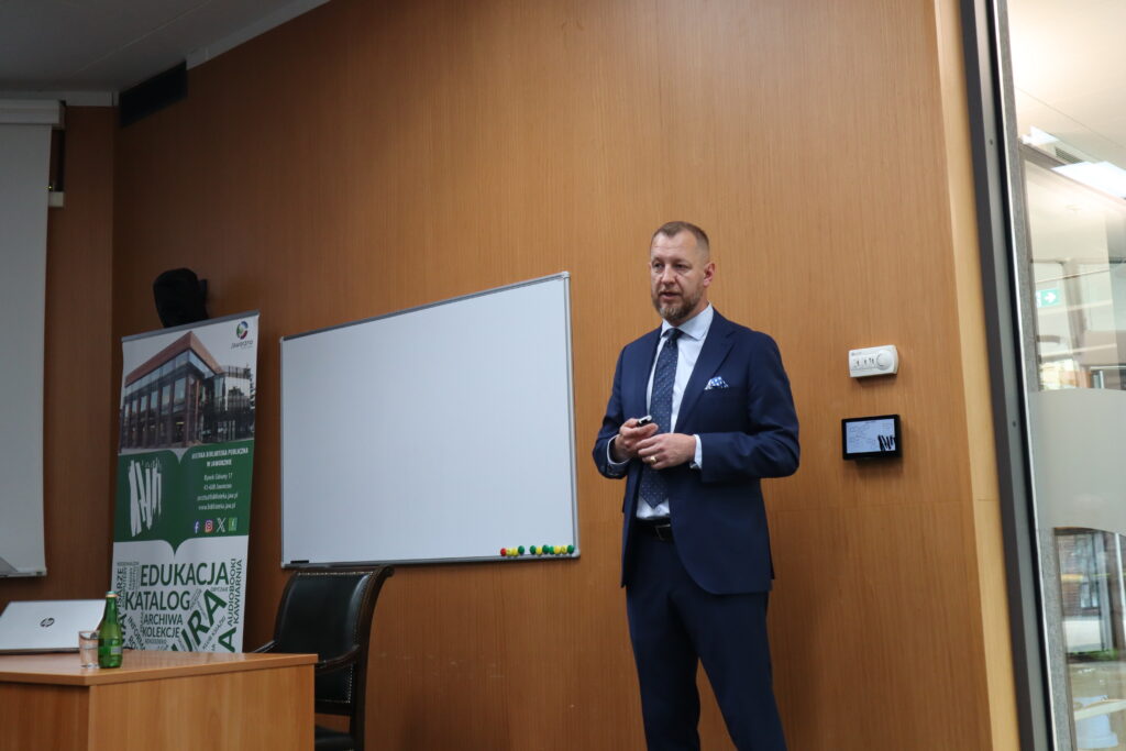 W audytorium, na tle rollupu biblioteki, stoi dr Piotr Uwijała. Komendant Miejski Policji w Jaworznie, ubrany w granatowy garnitur i białą koszulę, z poważną miną przemawia do niewidocznej publiczności.