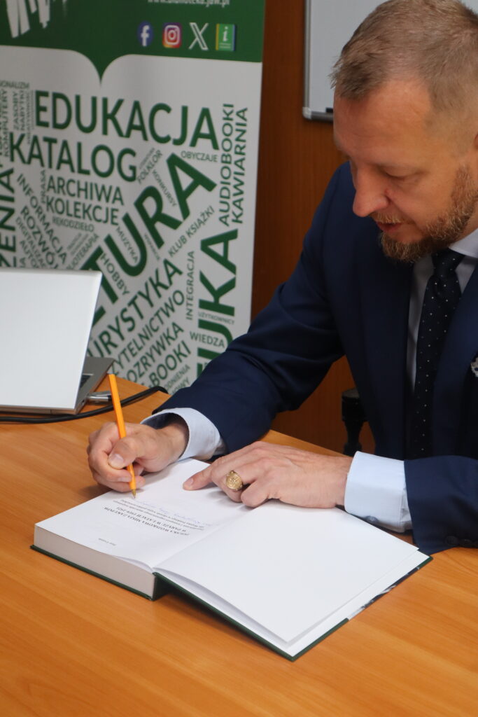 Zbliżenie na historyka, który siedzi przy biurku w audytorium i rozdaje autografy. Mężczyzna ma blond włosy i brodę, a ubrany jest w granatowy garnitur. W tle widać rollup biblioteki.