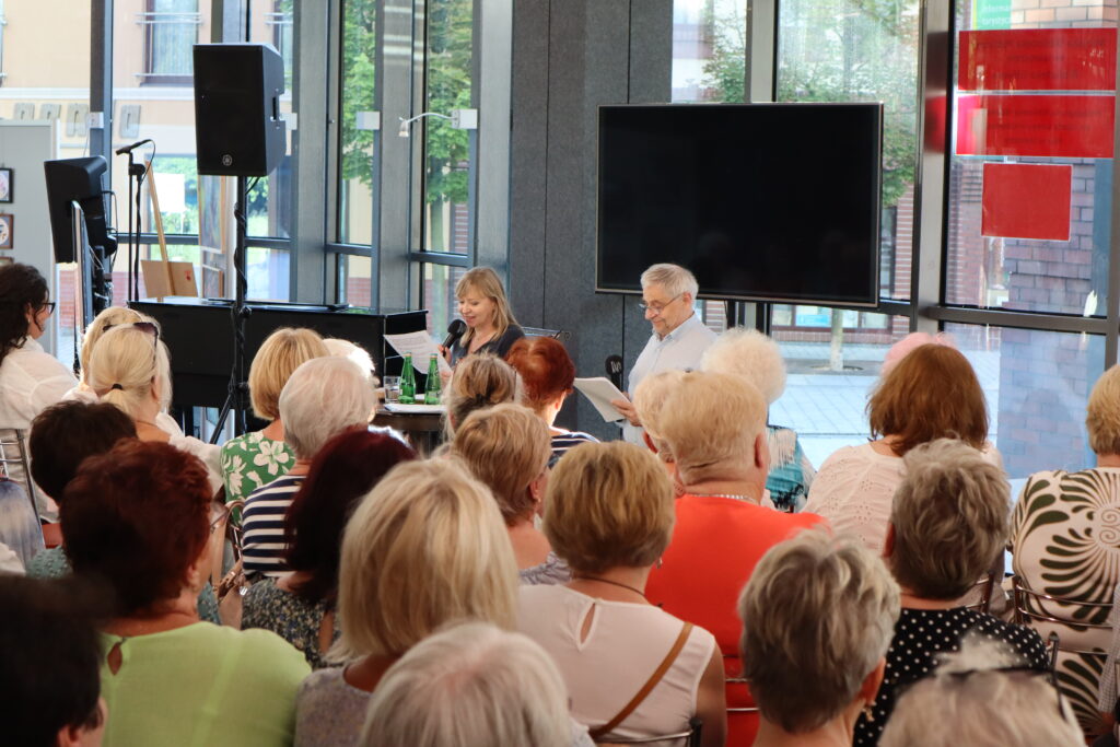 Hol Biblioteki Głównej na tle przeszklonej ściany siedzą przy okrągłym stoliku aktorzy - Dorota Segda i Jacek Romanowski. Blond włosa kobieta mówi do trzymanego w ręce mikrofonu. Na pierwszym planie widać siedzącą przed artystami publiczność.