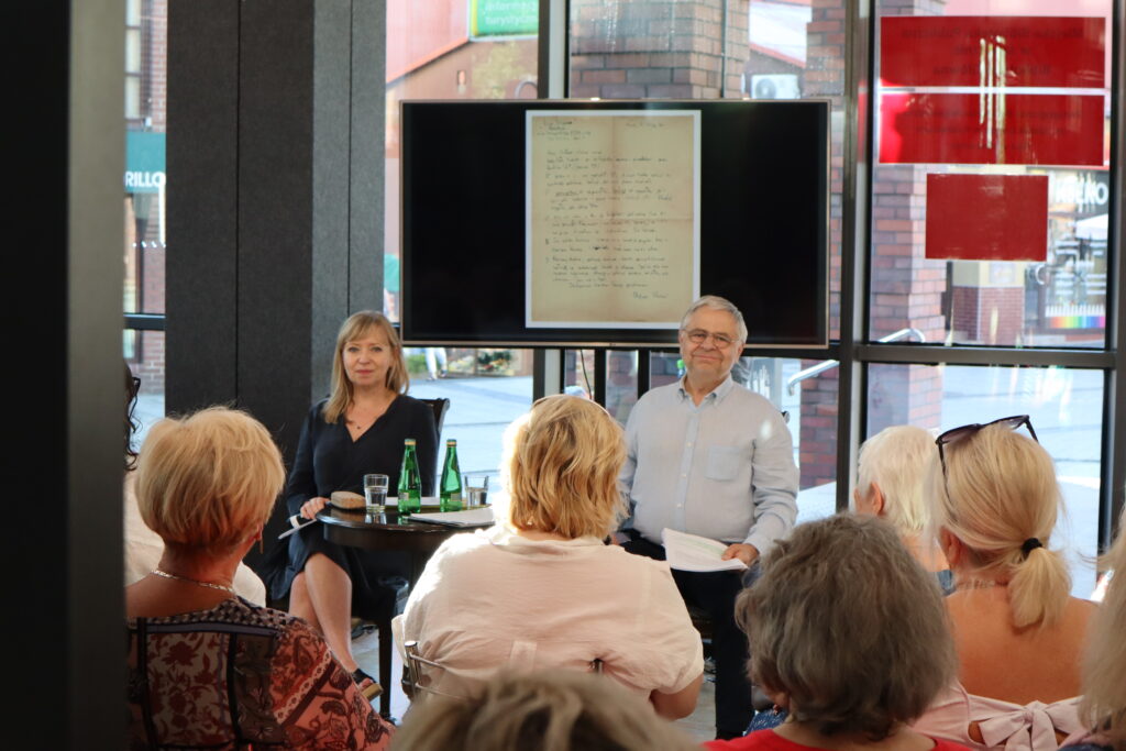 Hol Biblioteki Głównej na tle przeszklonej ściany siedzą przy okrągłym stoliku aktorzy - Dorota Segda i Jacek Romanowski. Za nimi na dużym telewizorze kadr prezentacji multimedialnej przedstawiający kopię odręcznego pisma - listu. Na pierwszym planie widać siedzącą przed artystami publiczność.