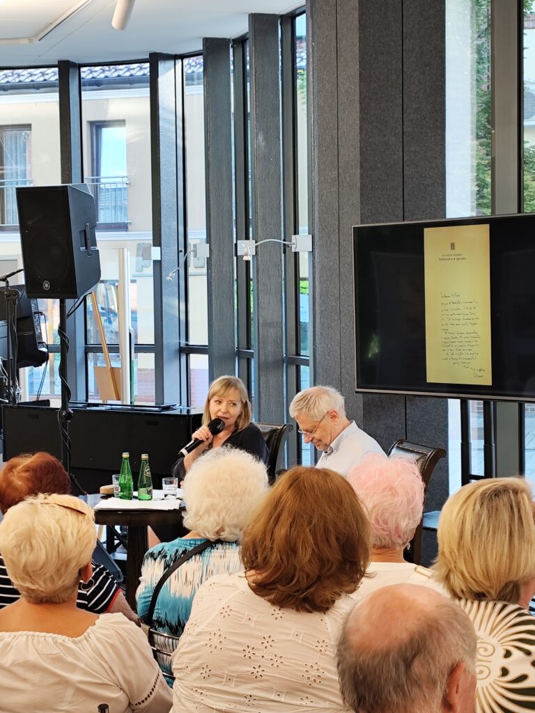 Hol Biblioteki Głównej na tle przeszklonej ściany siedzą przy okrągłym stoliku aktorzy - Dorota Segda i Jacek Romanowski. Za nimi na dużym telewizorze kadr prezentacji multimedialnej przedstawiający kopię odręcznego pisma - listu. Na pierwszym planie widać siedzącą przed artystami publiczność.