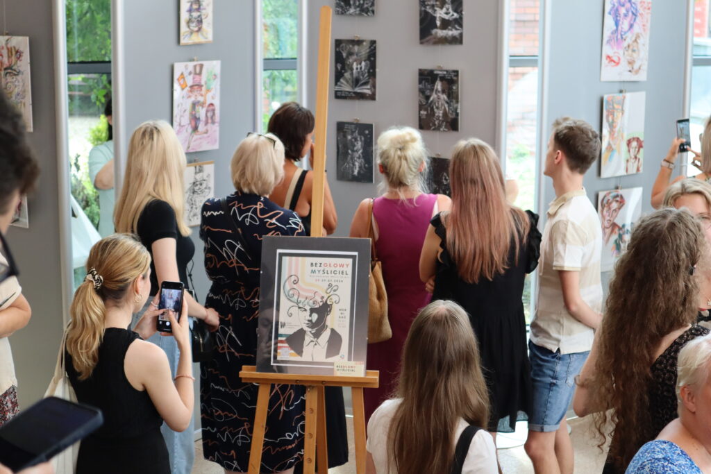 Zdjęcie przedstawia grupkę kilkunastu osób oglądających rysunki Marty Skubis, które rozwieszono na panelach wystawienniczych w bibliotecznej Galerii ExLibris.