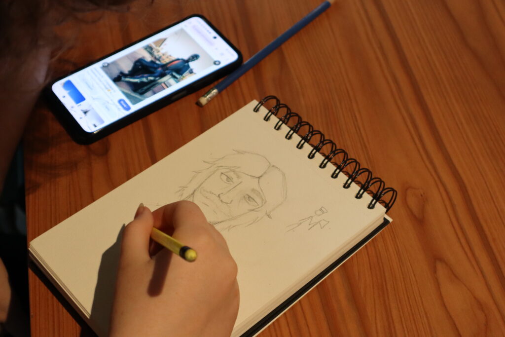 Zbliżenie na szkicownik, w którym jeden z uczestników tworzy postać komiksową inspirowaną wyglądem pomnika drwala z Jaworzna, którego zdjęcie wyświetla się na ekranie telefonu obok.