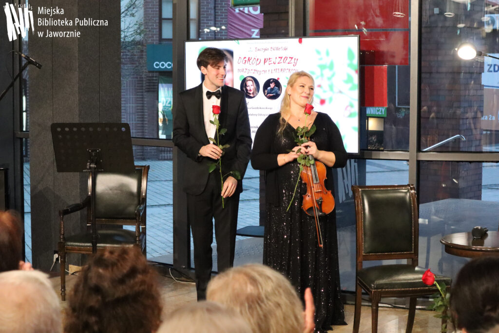 Na scenie w Bibliotece Głównej widzimy stojących z kwiatami artystów. Młody mężczyzna – pianista – ubrany jest w czarny garnitur, białą koszulę, przy szyi ma muszkę. Blondwłosa kobieta – skrzypaczka, ubrana w czarną cekinową sukienkę trzyma w rękach instrument – skrzypce. Tuż za nimi w tle widać telewizor, przedstawiający plakat koncertu i oszklone szyby Biblioteki Głównej.