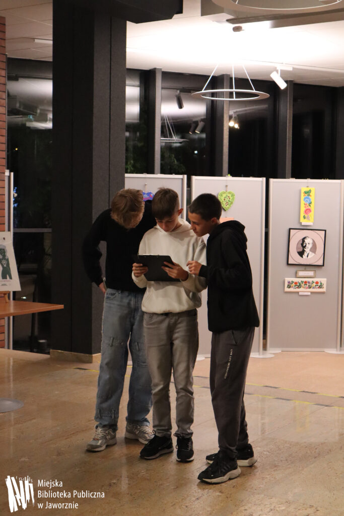 Na zdjęciu widzimy trzech chłopców, którzy stoją obok siebie i czytają jakąś kartkę. W tle widać biblioteczna Galerię ExLibris.