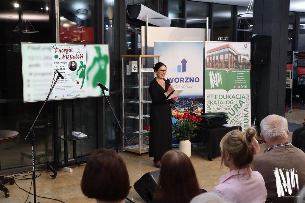 Zdjęcie przedstawia fragment holu Biblioteki Głównej w Jaworznie - to wysokie, przeszklone. Przez szyby widać, że na zewnątrz panuje mrok. Na scenie widzimy ciemnowłosą kobietę, ubraną w czarną sukienkę - to dyrektorka Biblioteki, która przemawia do mikrofonu. Z jej lewej strony odnajdziemy telewizor, na którym wyświetla się plakat projektu Energia Biblioteki. Z prawej strony umieszczono dwa roll-upy przedstawiające Bibliotekę Główną w Jaworznie oraz Jaworzno z lotu ptaka z dużym napisem Jaworzno poziom wyżej. Bliżej obiektywu dostrzeżemy kilka zwróconych w stronę sceny głów  - to uczestnicy wydarzenia. 