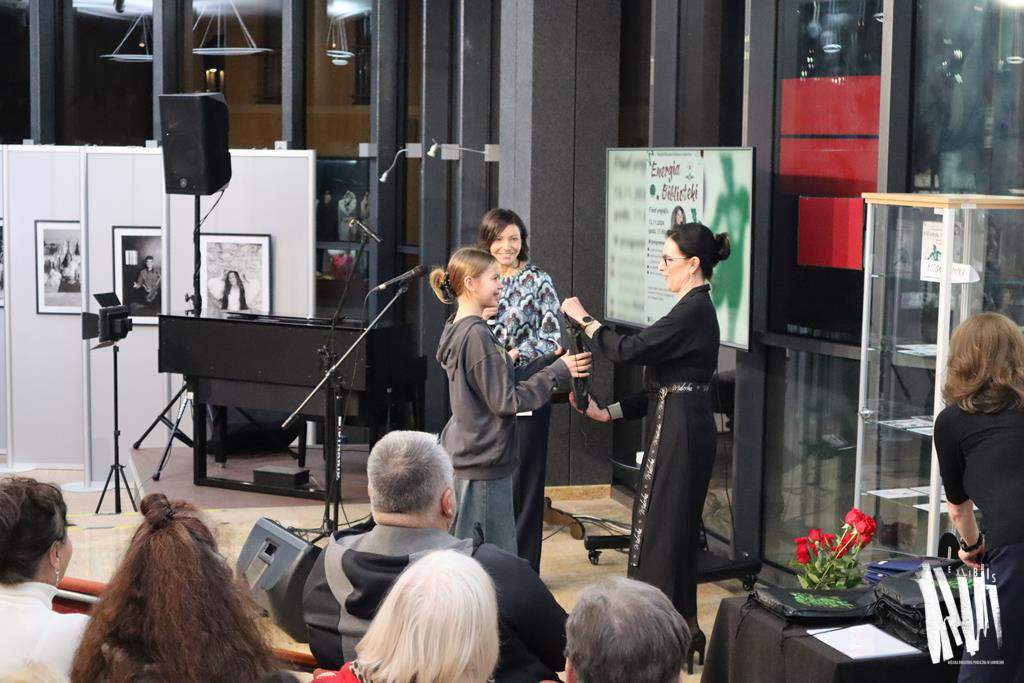 Zdjęcie przedstawia fragment holu Biblioteki Głównej w Jaworznie - to wysokie, przeszklone pomieszczenie. Przez szyby widać, że na zewnątrz panuje mrok. Na scenie widzimy ciemnowłosą kobietę, ubraną w czarną sukienkę - to dyrektorka Biblioteki, która wręcza materiałowy worek młodej dziewczynie w grafitowej bluzie i szerokich jeansach. Tuż za nią stoi kolejna kobieta, ciemnowłosa, ubrana we wzorzystą bluzkę. Za kobietami odnajdziemy telewizor, na którym wyświetla się plakat projektu Energia Biblioteki. Po lewej stronie zdjęcia dostrzeżemy niski, okryty ciemną płachta stolik, na którym ułożone są materiałowe worki koloru czarnego, z zielonym emblematem. Nad stolikiem pochyla się kobieta - ma długie, ciemne, pofalowane włosy, ubrana jest w czarną bluzkę i granatowe spodnie. Przy stoliku stoi wąska, oszklona gablota oraz czerwone róże. Bliżej obiektywu dostrzeżemy kilka zwróconych w stronę sceny głów  - to uczestnicy wydarzenia. 