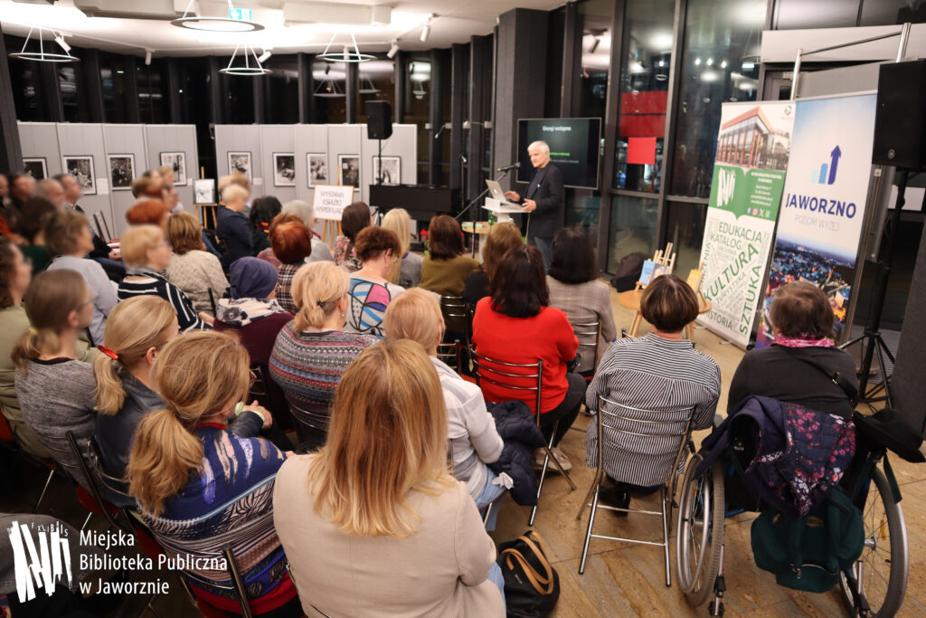 Widoczna część holu Biblioteki Głównej. Po prawej w tle przemawia Profesor Bogdan de Barbaro.Stoi za białą mównicą. Ma ciemne ubranie i białe, krótkie włosy. Mówi do licznie zgromadzonej przed nim widowni, na zdjęciu widać kilkadziesiąt osób siedzących bokiem i tyłem do aparatu, jedna z nich, po prawej stronie zdjęcia, jest na wózku inwalidzkim (kobieta). Po prawej widoczne są dwa banery: pierwszy reklamuje naszą instytucję, drugi miasto Jaworzno. W tle za wykładowcą slajd na dużym telewizorze, czerwone kwiaty na podłodze, stolik z wodą. Po lewej całkowicie z tyłu widoczny napis WYSTAWA KSIĄŻKI WSPIERAJĄCEJ, a całkiem z tyłu rozmyte czarno-białe zdjęcia. Za przeszklonymi ścianami mrok.