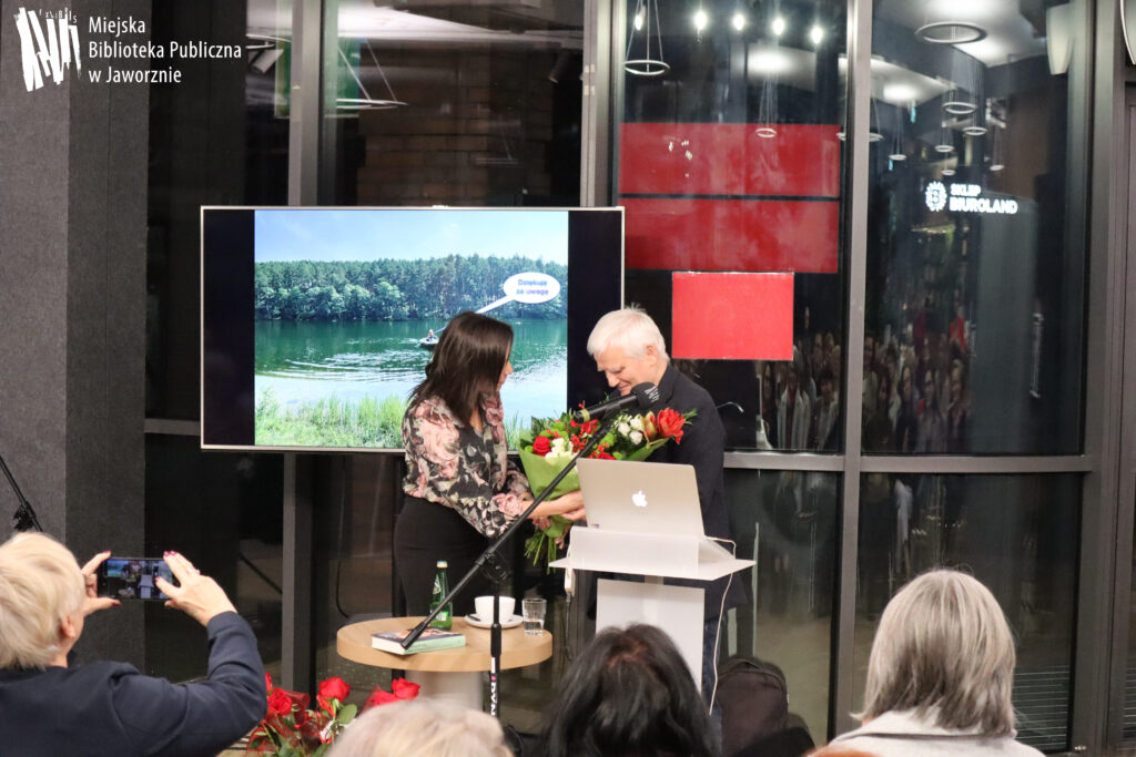 W centrum znajduje się Profesor de Barbaro i przedstawicielka jaworznickiej biblioteki. Kobieta ma ciemne średniej długości włosy, widoczna w lekkim profilu, ubrana w różowo-zielono-grafitową bluzkę i czarną spódnicę, wręcza Profesorowi duży bukiet kremowo-czerwonych kwiatów. Profesor stoi lekko pochylony nad bukietem. Ma ciemne ubranie i krótkie, białe włosy. Obok stolik z butelką wody, szklanką, filiżanką i książką. Z przodu widać fragment pierwszego rzędu widowni, tyłem, jedna z osób robi smartfonem zdjęcie. W tle czarny telewizor z kolorowym slajdem lasu, jeziora i łódki na jeziorze, z łódki komiksowy napis: Dziękuję za uwagę. Za przeszklonymi ścianami mrok i dwa czerwone prostokąty.