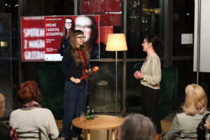 Na tle przeszklonej ściany holu Biblioteki Głównej stoją dwie kobiety - trzymająca w rękach herbacianą różę Magdalena Grzebałkowska i dyrektor Miejskiej Biblioteki Publicznej w Jaworznie - Monika Rejdych, ciemnowłosa kobieta ubrana jest w dzianinową bluzkę ze złotą nitką i czarną, długą spódnicę. Włosy ma spięte. Panie uśmiechają się do siebie.