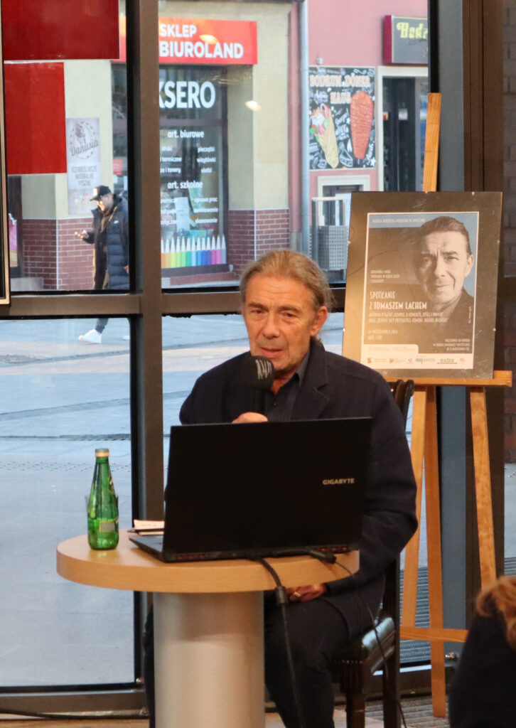 Przed oszkloną ścianą holu Biblioteki Głównej, siedzi przy okrągłym stoliku mężczyzna - Tomasz Lach, ma szpakowate włosy, spięte w mały kucyk z tyłu głowy. Ubrany jest w granatową marynarkę, ciemną koszulę i czarne spodnie. W ręce trzyma mikrofon bezprzewodowy. Na stoliku znajduje się otwarty laptop. Po prawej stronie za mężczyzną stoi sztaluga, na której jest oprawiony w ramę plakat zapowiadający spotkanie z jego wizerunkiem. Po lewej stronie za mężczyzną jest telewizor na stojaku, na którego ekranie widać zdjęcie Krzysztofa Komedy i Marka Hłaski. Na pierwszym planie widoczne są osoby siedzące na krzesłach - uczestnicy spotkania.