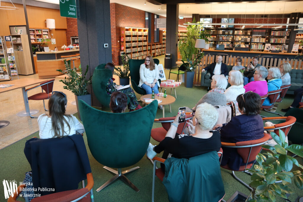 Zdjęcie przedstawia fragment wnętrza Biblioteki Głównej w Jaworznie. W oddali widać kilka regałów wypełnionych książkami, stanowisko obsługi czytelników w Wypożyczalni dla Dorosłych oraz Bibliotecafe. Zdjęcie wykonano w strefie seniora. Na dużych, zielonych fotelach siedzą dwie kobiety. Pierwsza od prawej to gość spotkania - Patrycja Sawiuk. Ubrana jest niebieskie jeansy i biała marynarkę. Ma długie, kasztanowe włosy, na nosie okulary. Trzyma ręce splecione przed sobą. Obok niej widzimy bibliotekarkę, która patrzy na pisarkę. Ma ciemne włosy i sukienkę w podobnej tonacji z kolorowymi motywami kwiatowymi. Tuz przed nimi stoi niski stolik, na którym dostrzeżemy dwie butelki wody mineralnej, szklanki i książki. Przy autorce od prawej strony stoi podobny stolik z książkami i plakatem promującym spotkanie. Bliżej obiektywu widzimy publiczność - to uczestnicy spotkania.