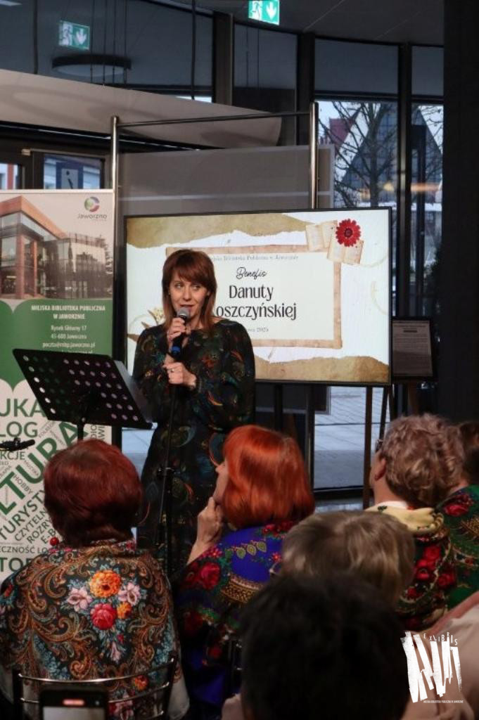 Przy wejściu do Biblioteki Głównej, na tle dużego ekranu prezentującego slajd prezentacji, w tym napis: Benefis Danuty Noszczyńskiej, stoi przy mikrofonie na statywie rudowłosa kobieta, ubrana w sukienkę we wzory przypominające pawie pióra, przed nią stoi czarny pulpit. Na pierwszym planie widoczna jest siedząca publiczność. Panie w pierwszym rzędzie mają na sobie ludowe chusty.