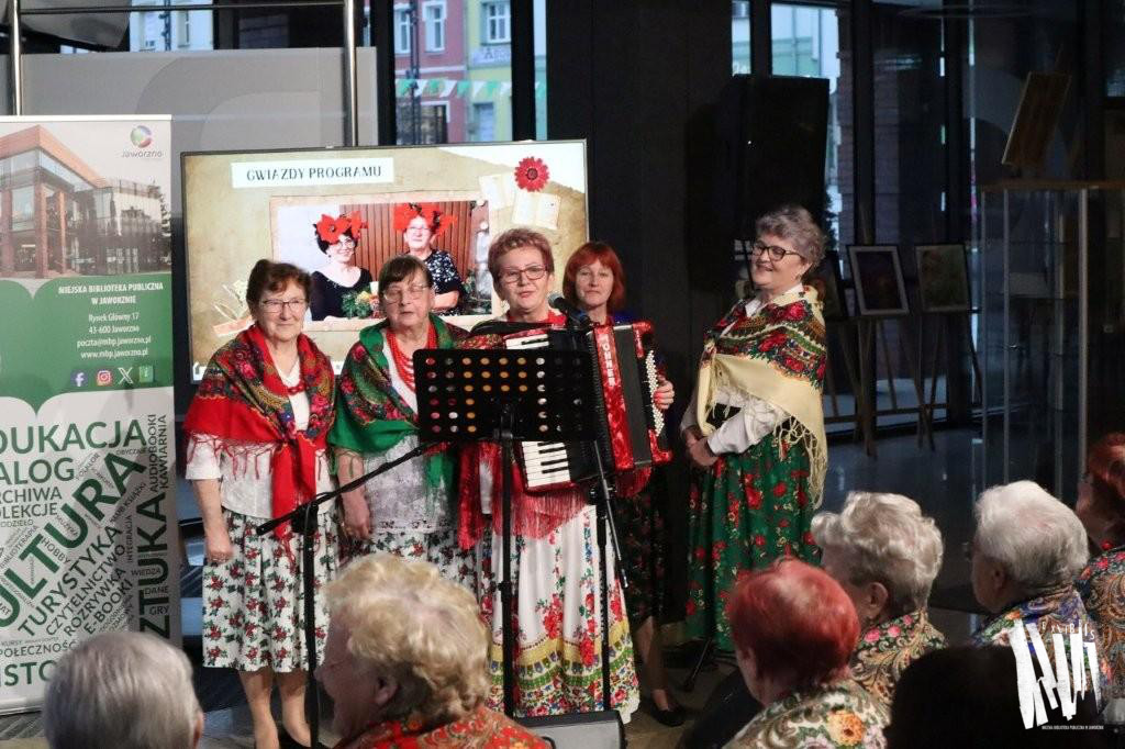 Przed przeszkloną ścianą Biblioteki Głównej, na tle baneru informującego o działalności MBP w Jaworznie i ekranu telewizora z wyświetlonym kadrem prezentacji multimedialnej, stoi przy mikrofonie na statywie pięć kobiet w strojach ludowych. Jedna z nich gra na akordeonie i śpiewa. Na pierwszym planie widoczna siedząca w rzędzie publiczność.