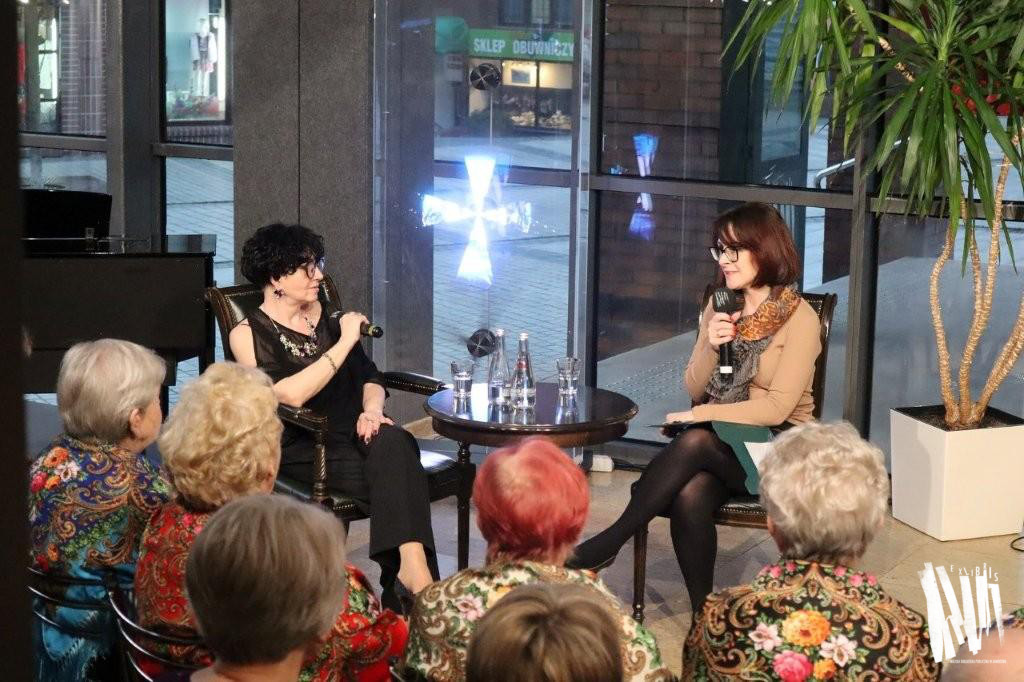 Przed przeszkloną ścianą Biblioteki Głównej, przy okrągłym stoliku siedzą i rozmawiają dwie kobiety, obie trzymają w dłoniach mikrofony. Jedna, ubrana jest w czarne spodnie i czarną bluzkę z odkrytym ramieniem. Druga ma na sobie beżowy sweterek, zieloną krótką spódnicę i związany pod szyją szal z panterkowym wzorem. Obie noszą okulary. Przed nimi siedzi publiczność.