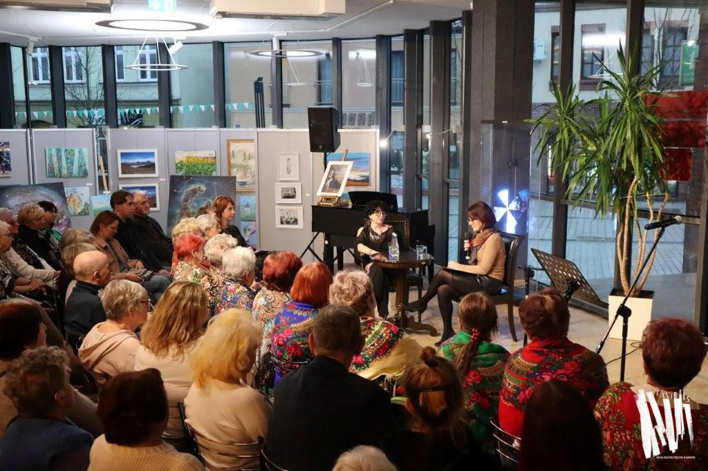Przed przeszkloną ścianą Biblioteki Głównej, przy okrągłym stoliku siedzą dwie kobiety. Jedna, ubrana jest w czarne spodnie i czarną bluzkę z odkrytym ramieniem. Druga ma na sobie beżowy sweterek, zieloną krótką spódnicę i związany pod szyją szal z panterkowym wzorem, trzyma w ręce mikrofon. Obie noszą okulary. Przed nimi siedzi publiczność.