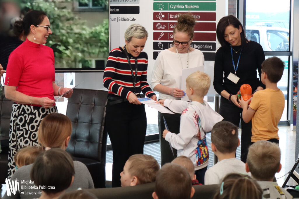 Zdjęcie przestawia cztery kobiety, przed którymi stoi dwójka chłopców. Dzieci wręczają kobietom po kolei podziękowania za udział w spotkaniu w formie dyplomu oraz pomarańczową bombonierkę w kształcie pluszowego misia. W tle po lewej stoi telewizor, po prawej tablica informacyjna. Na pierwszym pranie znajdują się dzieci odwrócone plecami do zdjęcia.
